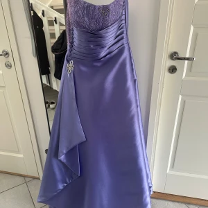 Lila balklänning från Jovani - Extremt snygg off shoulder balklänning som är gjord av satin, den har jätte fina stenar sydda vid bröstet och har till och med en matchande sjal till! Är 170cm lång och klänningen nådde marken perfekt med klackar på. Hör gärna av för bilder på sjalen. Klänningen är storlek 10 US men är insydd så till den som är storlek 36/34 sitter den perfekt. Pris går även att förhandlas vid snabbt köp!