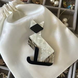 kattbrosch i svart och vitt - Unik brosch i form av en stiliserad katt med geometriska former. Broschen har svart huvud och svans, vit kropp med marmorerad effekt och detaljer i silver. Perfekt accessoar för att ge din outfit en cool och lekfull touch.