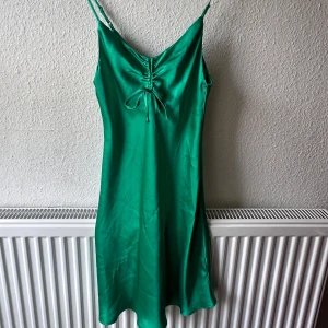 Grön satin klänning från Bik Bok - Supersnygg grön klänning från Bik Bok i glansigt satinmaterial. Den har smala axelband, v-ringning och en söt snörningsdetalj framtill. Modellen är kort och har en lätt figurnära passform. Liten skada framtill, syns inte så mycket.