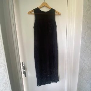 Svart spetsklänning med mesh och dragkedja - Snygg svart långklänning med spetsdetaljer och transparent mesh upptill. Klänningen är ärmlös och har en guldig dragkedja i ryggen som ger en cool kontrast. Perfekt för dig som gillar stilrena och eleganta plagg med det lilla extra.