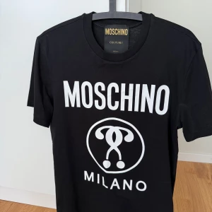 Svart Moschino t-shirt  -  svart t-shirt från Moschino med stort vitt logotryck på bröstet och Milano-text. Klassisk rund hals och korta ärmar. Tillverkad i 100% bomull för en skön känsla. Perfekt för dig som gillar statement-plagg och streetstyle. I princip oanvänd vilket också syns på skicket. Äkta och orderbekräftelse finns till hands! Hör av er vid frågor och funderingar, mvh 