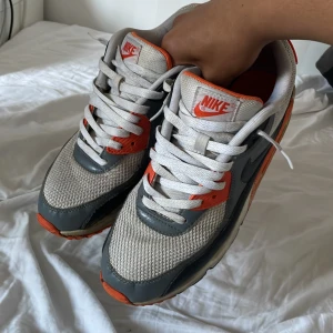 Nike Air Max 90 grå och orange - Nike Air Max 90 sneakers i grått, vitt och orange med klassisk Air Max-dämpning. Ovandel i mesh och skinn, snygga detaljer och tydlig Nike-logga på hälen. Perfekta för dig som gillar retrostil och vill sticka ut med färg.