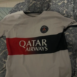 PSG Mbappé fotbollströja vit/röd - Snygg PSG fotbollströja med Mbappé och nummer 7 på ryggen. Tröjan är vit med breda röda och svarta horisontella ränder över bröstet, Qatar Airways-logga och PSG-emblem. Kortärmad modell i lätt och ventilerande material, perfekt för match eller träning.