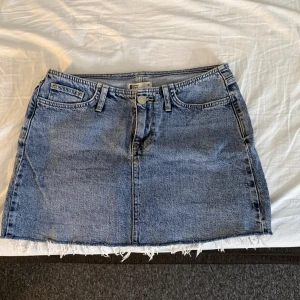 Blå jeanskjol med fransig kant - Snygg blå jeanskjol från Gina Tricot med klassisk femficksdesign och fransig, rå kant nertill. Kjolen har bälteshällor och dragkedja med knapp framtill. Perfekt för en avslappnad och trendig look.