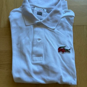 Vit Lacoste x La Casa de Papel pikétröja - Vit pikétröja från Lacoste i samarbete med La Casa de Papel. Klassisk krage, knäppning framtill och broderad krokodil-logga i rött och grönt på bröstet samt original-logga i nacken. Tillverkad i bomull med klassisk fit. (Storlek 5 = large.)