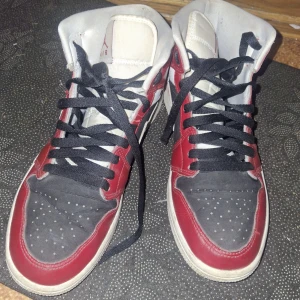 Nike Air Jordan 1 Chicago sneakers - Säljer ett par Nike Air Jordan 1 Chicago sneakers med klassisk färgkombination i rött, svart och vitt. Skorna har svarta snören, vit sula och ikonisk Swoosh-logga på sidan. Tillverkade i skinn och syntet, med perforerad tå och högt skaft för en sportig look.