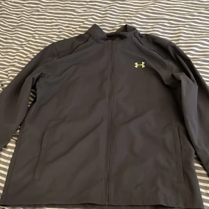 Mörkgrå vindjacka från Under Armour - Snygg svart vindjacka från Under Armour med limegrön logga på bröstet och stort tryck på ryggen. Jackan har hel dragkedja, två fickor och meshfoder för extra komfort. Perfekt för träning eller när du vill ha en sportig look.