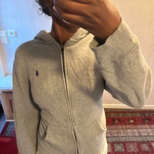 Grå hoodie från Ralph Lauren - Snygg grå hoodie från Ralph Lauren med dragkedja framtill och klassisk broderad logga på bröstet. Tröjan har huva, fickor och är tillverkad i mjuk bomull. Perfekt för en chill och avslappnad stil.