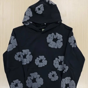 Svart hoodie från demim tears M - Cool svart hoodie från denim tears med stora gråa blommönster över hela tröjan. Tröjan har huva och lång ärm, perfekt för dig som gillar streetwear med unik design. Tillverkad i mjuk bomull för skön känsla.