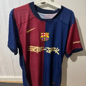 FC Barcelona Lamine Yamal Nike tröja - Snygg FC Barcelona matchtröja från Nike med Lamine Yamal och nummer 19 på ryggen. Tröjan har klassiska blå och röda paneler, gul Nike-logga och klubbmärke framtill samt Cactus Jack-tryck. Tillverkad i lätt och ventilerande material, perfekt för fotbollsälskare.