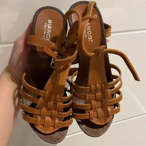 Bruna klackskor från Bianco - Säljer ett par bruna klackar från Bianco med hög klack och platåsula. Skorna har flera remmar över foten, spänne vid ankeln och dekorativa nitar längs sidorna. Perfekta för dig som vill ha en snygg och stabil klacksko i skinnimitation.