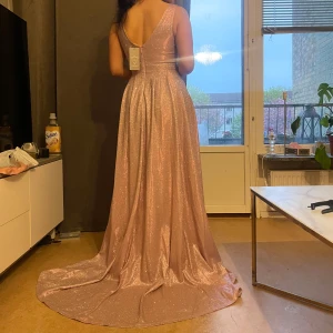 Glittrig ljusrosa långklänning med v-ringning - Säljer en fantastisk glittrig ljusrosa långklänning med djup v-ringning och öppen rygg. Klänningen har ett elegant fall och är ärmlös, perfekt för bal eller fest. Materialet skimrar vackert i ljuset och ger en lyxig känsla.