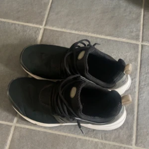 Svarta Nike Presto sneakers - Säljer ett par svarta Nike Presto sneakers med vit sula och svarta skosnören. Skorna har en sportig design med rund tå och meshdetaljer för extra komfort. Perfekta för dig som gillar stilrena och bekväma sneakers.