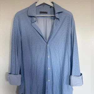 Ljusblå rutig skjorta från Corneliani - Snygg ljusblå skjorta från Corneliani med diskret rutigt mönster. Klassisk krage och knäppning framtill. Skrynkelfri så behöver inte strykas. Perfekt för en stilren look till sommaren. +2000kr nypris, nyskick!