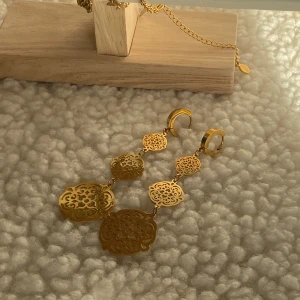 Guldiga hängande örhängen med utskurna mönster - Säljer ett par statement örhängen i guld med flera utskurna, dekorativa plattor i olika former. Örhängena är långa och har en elegant, bohemisk känsla. Perfekta för att lyfta din look med lite extra bling. Rostfritt stål/guldpläterad 