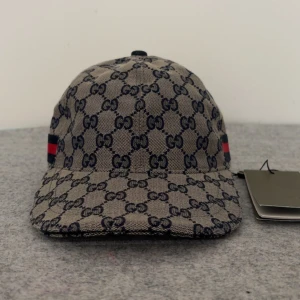 Gucci keps beige - Skicka  dm för prisförslag eller video. Storlek ”M”  Säljer en beige keps från Gucci med klassiskt GG-monogram i mörkblått. Kepsen har justerbart svart läderband bak och röd-blå randig detalj på sidan. Perfekt accessoar för en trendig look.