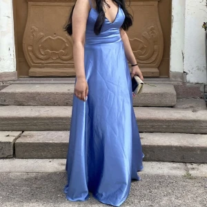 Balklänning - Säljer en elegant ljusblå långklänning med djup v-ringning och breda axelband. Klänningen har en glansig finish och markerad midja, perfekt för speciella tillfällen. Stilren och klassisk design.