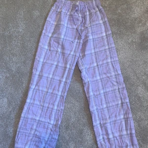 Lila rutiga byxor från Gina Tricot - Lila och vita rutiga byxor från Gina Tricot med resår i midjan och snörning. Byxorna har en avslappnad passform och är perfekta för en chill stil. Mjukt bomullsmaterial som känns skönt mot huden.