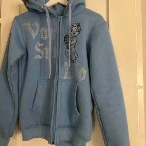 Ljusblå hoodie med dragkedja och strasstext - Säljer en ljusblå hoodie från Von Dutch med dragkedja, huva och fickor framtill. Denna färg sälj inte längre.