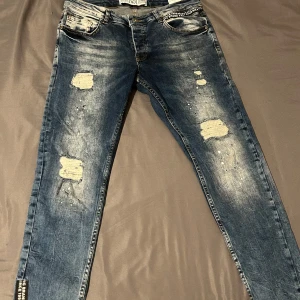 Blå slitna jeans från VSCT - Säljer ett par blå jeans från VSCT med slitna detaljer och broderad logga på bakfickan. Jeansen har normal passform och klassisk femficksdesign. Perfekta för dig som gillar en lite ruffare streetstil.