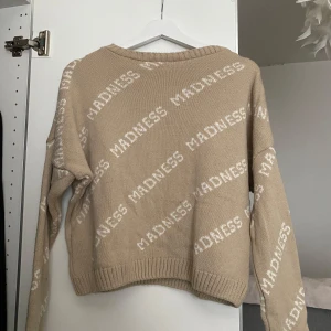 Beige stickad tröja från Madlady med textmönster - Säljer en beige stickad tröja från Madlady med vit text 'MADNESS' över hela tröjan. Tröjan har rund halsringning, långa ärmar och ribbade muddar. Perfekt för dig som gillar statement-plagg och vill ha något mysigt och trendigt. Den är knappt använd, i väldigt fint skick! Pris kan diskuteras vid snabb affär!!