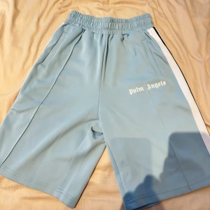 Ljusblå shorts från Palm Angels - Klassiska Palm Angels shorts i ljusblått med vit sidoremsa och logga tryckt på vänster ben. Storlek L, felfria som nyttskick. Säljs för att de inte passar mig. Pris kan diskuteras
