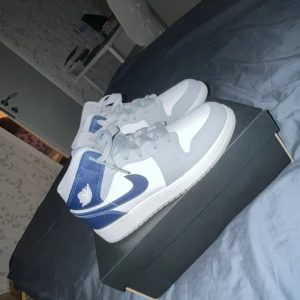 Nike Air Jordans Mid i vitt, grått och blått - Säljer ett par Nike Air Jordans Mid med vit bas, gråa paneler och blå detaljer. Klassisk Jordan-logga på sidan och blå swoosh. Skorna har snörning och är perfekta för dig som gillar streetwear och sneakers.