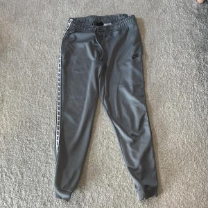 Gråa byxor Nike  - Grå träningsbyxor från Nike med svarta loggor längs sidorna. Byxorna har smal passform, muddar vid bensluten och en bakficka. Perfekta för sport eller chill.