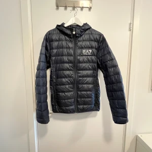 Svart pufferjacka från EA7 Emporio Armani - Säljer en svart pufferjacka från EA7 Emporio Armani med huva och dragkedja. Jackan har quiltad design och logga på bröstet. Perfekt för kyliga dagar och har en sportig look. 