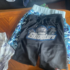 Svarta shorts från Trapstar London Shooters - Svarta shorts med blå och grå tryck från Trapstar London Shooters. De har snörning i midjan och ett stort grafiskt tryck framtill. Perfekta för en avslappnad streetwear-look.