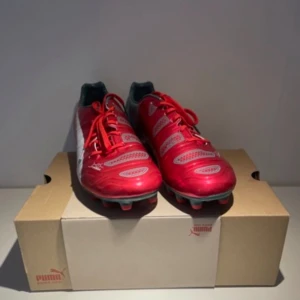Röda och vita fotbollsskor från Puma - Säljer ett par snygga fotbollsskor från Puma i rött, vitt och ljusblått. Skorna har snörning och är designade för spel på gräs med fasta dobbar. Perfekta för dig som vill sticka ut på planen!