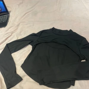 Svart långärmad croppad topp - Säljer en svart långärmad croppad topp med hög hals och unik cut-out detalj över bröstet. Perfekt för att skapa en trendig och edgy look. Materialet är ribbat och stretchigt för en bekväm passform.