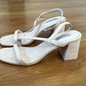 Beige sandaler med klack - Säljer ett par stilrena beige sandaler  med bred klack och smala remmar. Skorna har öppen tå och är tillverkade i mocka. Helt oanvända 