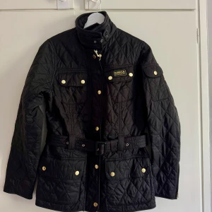 Svart quiltad jacka från Barbour International - Säljer en svart quiltad jacka från Barbour International med bälte i midjan och guldiga knappar. Jackan har hög krage, lång ärm och flera fickor framtill. Klassisk Barbour-logga på bröstet. Använd ett fåtal gånger, i jättefint skick!!