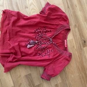 Mönstrad blus från Indiska - Säljer en mörk rosa blus med vackra broderade detaljer och tofsar framtill. Blusen har korta ärmar med smock och dekorativt mönster vid halsringningen. Perfekt för dig som gillar bohemisk stil.