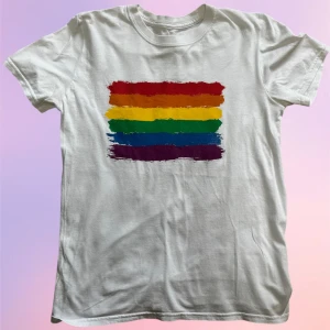 Vit t-shirt med regnbågsflagga - Vit t-shirt med Pride flagga! Bra kvalitet! Skulle gissa att tröjan är en 100% bomullströja men tröjan har inga lappar alls. Skulle säga att tröjan är en storlek S/M. 