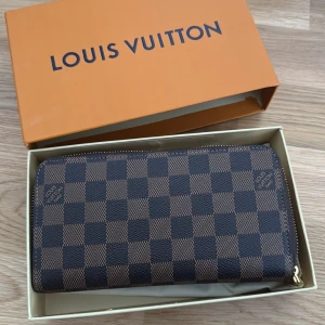 Superfin plånbok  - Säljer en klassisk plånbok i brunt och beige med rutigt mönster och guldiga dragkedjedetaljer. Insidan är ljusrosa med flera kortfack och fack för sedlar och mynt. Perfekt för dig som vill ha en lyxig accessoar.