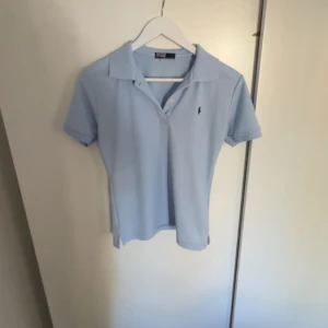 Ljusblå pikétröja från Ralph Lauren - Säljer en klassisk ljusblå pikétröja från Ralph Lauren med korta ärmar och krage. Tröjan har en liten broderad logga på bröstet och är tillverkad i mjukt bomullsmaterial. Perfekt för en stilren och avslappnad look.