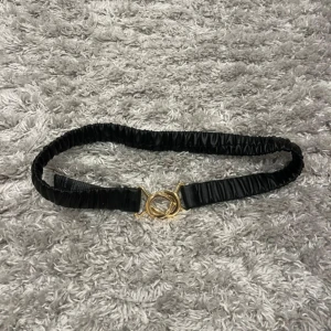 Svart resårbälte med guldfärgat spänne - Säljer ett svart resårbälte från Gina Tricot med snyggt guldfärgat spänne framtill. Bältet har en rynkad design och är elastiskt för en bekväm passform. Perfekt accessoar för att lyfta din outfit.