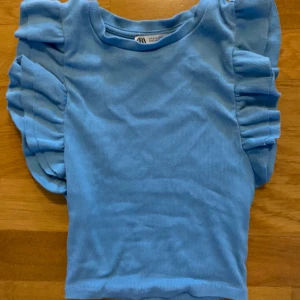 Blå topp med volangärmar  - Supersöt ljusblå topp från Zara med volangdetaljer på axlarna. Toppen har rund halsringning och är tillverkad i ett mjukt, ribbat material. Perfekt för att addera lite extra till din outfit!