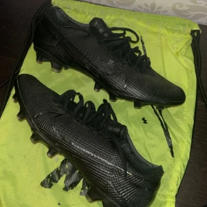 Svarta Nike Mercurial fotbollsskor - Säljer ett par svarta Nike Mercurial fotbollsskor med texturerad yta och snörning. Skorna har rund tå och platt sula med dobbar för bra grepp på planen. Perfekta för dig som vill ha stilrena och snabba skor på matchen.