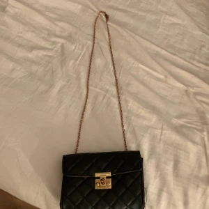 Svart quiltad axelväska med guldkedja - Säljer en svart quiltad axelväska med guldfärgad kedja och snyggt guldfärgat lås framtill. Väskan har klassisk design och är perfekt för att lyfta din outfit. Rutmönstrad yta ger en lyxig känsla. Passar till många olika stilar.