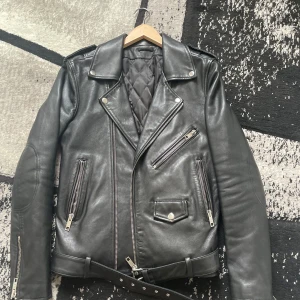 Svart skinnjacka med bälte och dragkedjor - Säljer en klassisk svart skinnjacka i bikerstil med flera dragkedjor, snedställd front, axelklaffar och bälte i midjan. Jackan har quiltat foder och detaljer som fickor med dragkedja. Denna jackan är av märket BLK DNM och den är köpt för 8,500. Jackan är av lamb skin och är av hög kvalitet 