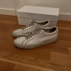 Vita sneakers från Common Projects - Säljer ett par klassiska vita sneakers från Common, Skorna har rund tå, platt sula och snörning. Perfekta för en stilren och minimalistisk look. Skokartong och extra skosnören medföljer.