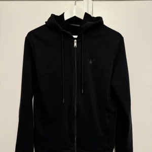 Svart hoodie från Emporio Armani - Säljer en stilren svart hoodie från Emporio Armani. Den har en dragkedja framtill och en diskret logga på bröstet. Perfekt för både vardag och chill. Passar till allt! Storlek M