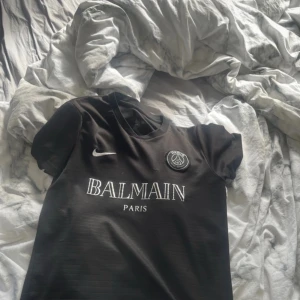 Svart PSG fotbollströja x Balmain - Snygg svart PSG fotbollströja i samarbete med Balmain. Tröjan har korta ärmar, rund hals och tydliga vita tryck med Nike-logga, Balmain Paris och PSG-emblem på bröstet. Materialet är lätt och andas, perfekt för fotboll eller streetwear.