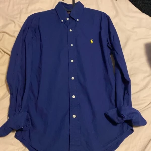 Ralph lauren skjorta  - Klassisk blå skjorta från Polo Ralph Lauren i slim fit-modell. Skjortan har långa ärmar, knappar framtill och den ikoniska broderade loggan på bröstet. Perfekt för en stilren och tidlös look.