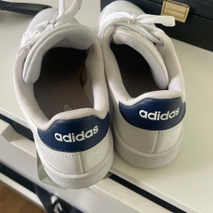 Vita Adidas sneakers med blå detalj - Säljer ett par klassiska vita Adidas sneakers med blå detalj på hälen och logga. Skorna har snörning, perforerade sidor och platt sula. 🥹