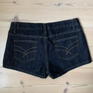 Mörkblå jeansshorts  - Snygga mörkblå jeansshorts med klassisk femficksdesign och kontrastsömmar. Modellen är kort och har normal passform med knapp- och dragkedjestängning framtill. Köpt vintage. 
