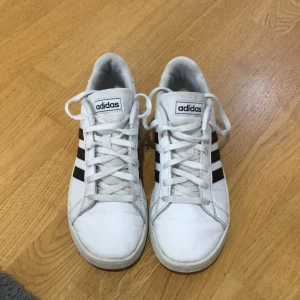 Vita Adidas sneakers med svarta ränder - Klassiska vita Adidas sneakers med svarta ränder på sidorna och vit sula. Skorna har snörning och är tillverkade i syntetmaterial. Perfekta för en sportig och stilren look.